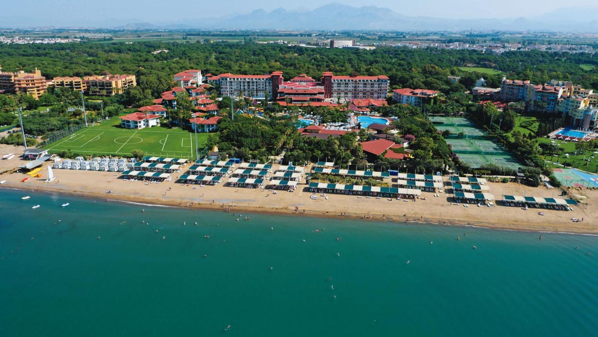 Belconti Resort Hotel panorāmas skats no jūras puses — viesnīcas korpusi, futbola laukums, tenisa korti un plaša pludmales josla