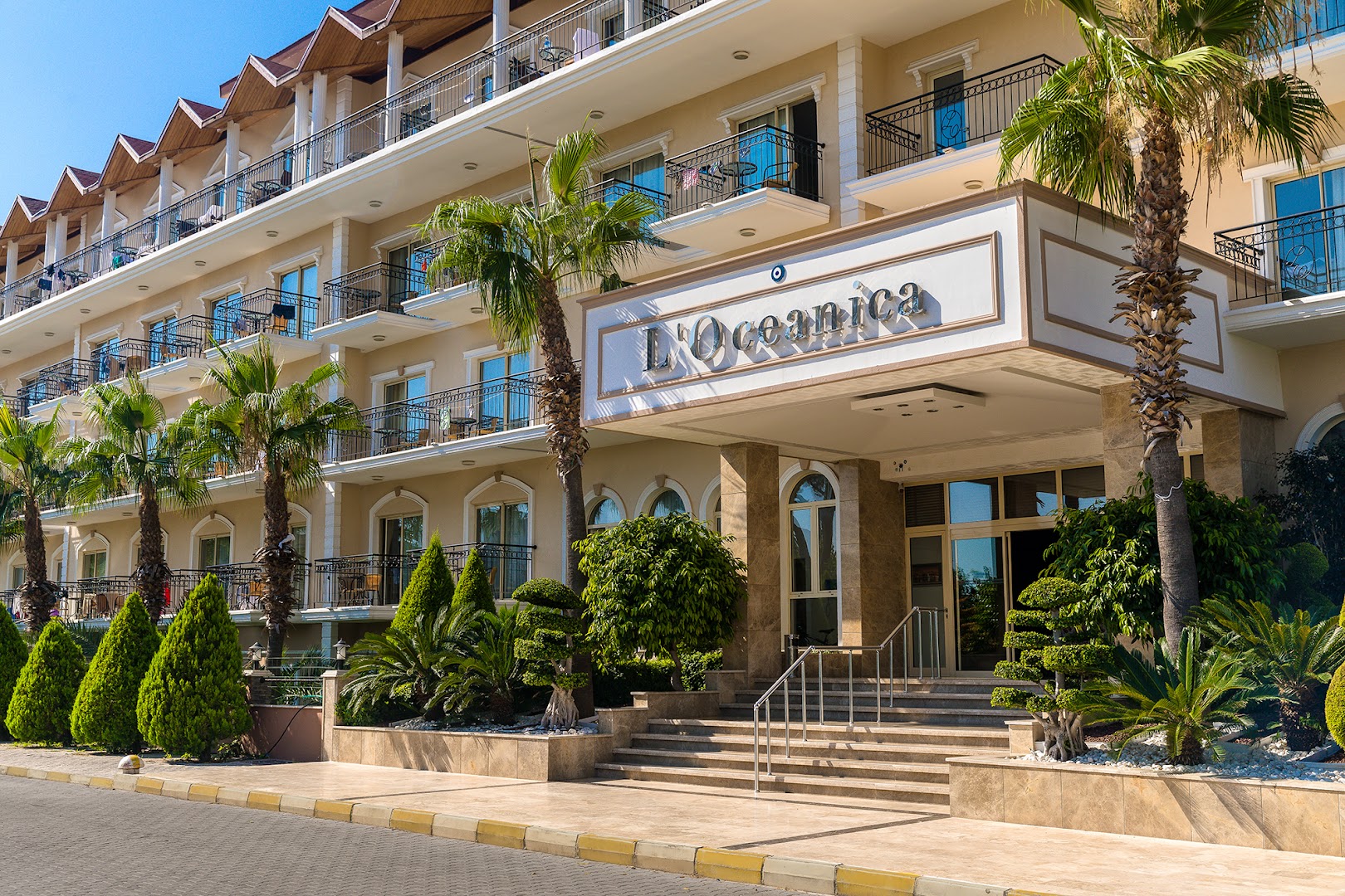 L'oceanica Beach Resort Hotel galvenā ieeja un fasāde ar palmām, dekoratīviem krūmiem un numuru balkoniem saulainā dienā