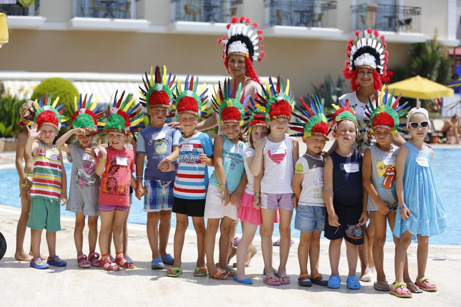 Bērnu izklaides pasākums pie L'oceanica Beach Resort Hotel baseina — animatori un bērni krāsainos tērpos ar spalvu galvas rotām