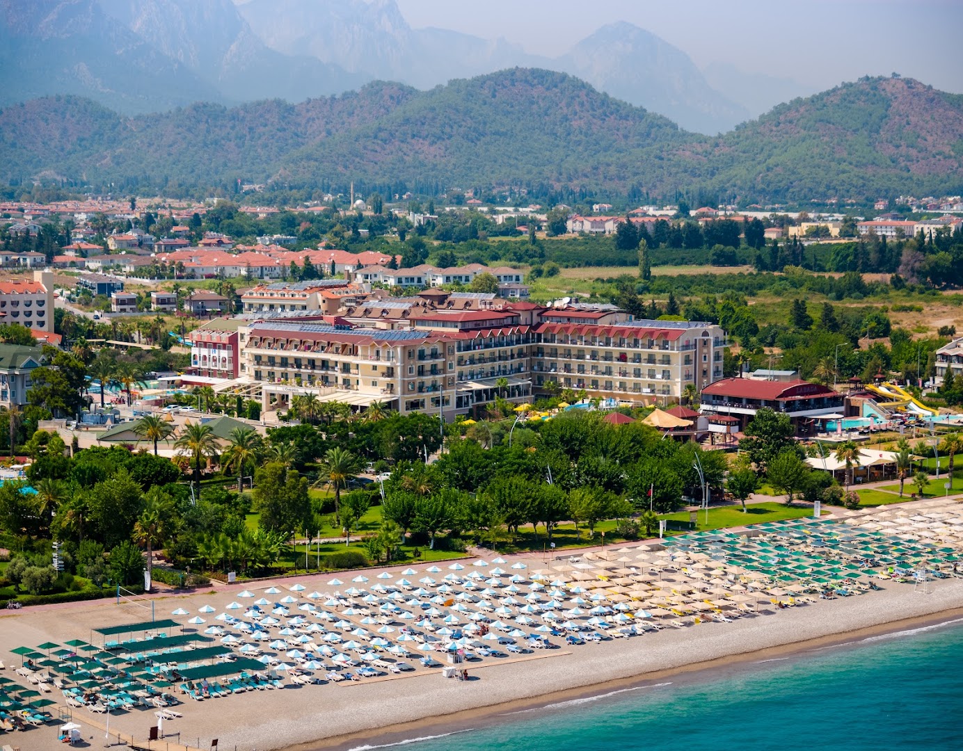 L'oceanica Beach Resort Hotel panorāmas skats Kemeras reģionā — plaša oļu pludmale ar saulessargiem, viesnīcas ēka un kalni fonā