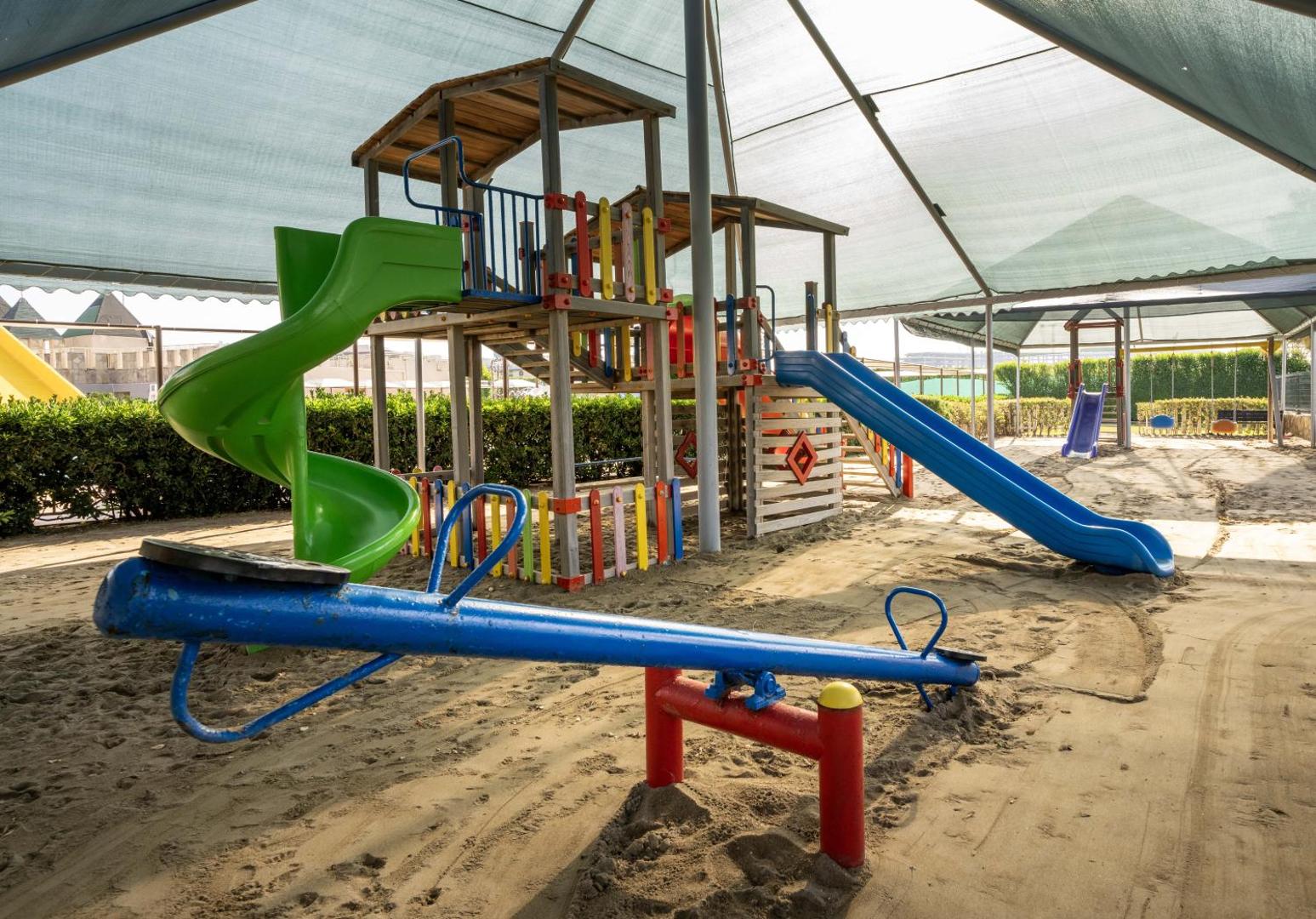 Kaya Belek bērnu rotaļu laukums smiltīs zem aizsargājošas nojumes ar slidkalniņiem un šūpolēm.