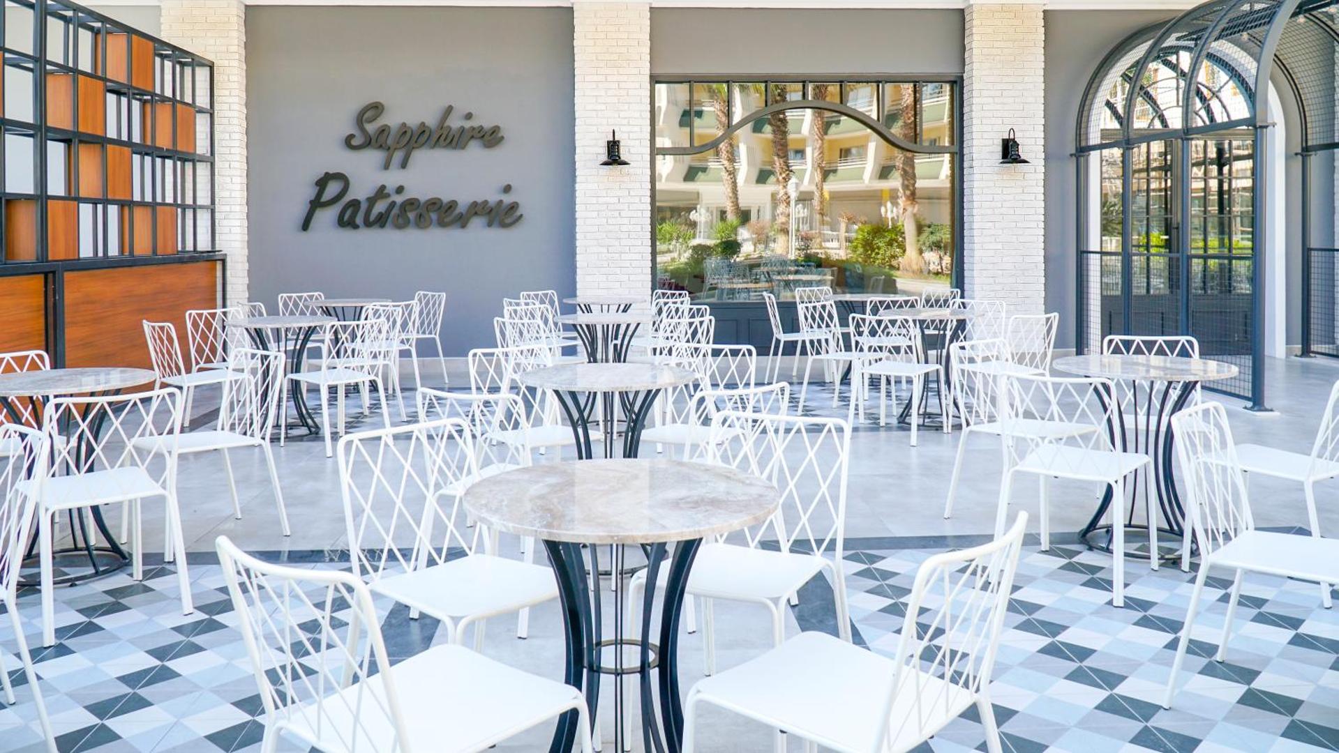 Sapphire Patisserie āra terase viesnīcā Rai Premium Tekirova ar baltiem metāla krēsliem un marmora galdiņiem uz rakstainas flīžu grīdas