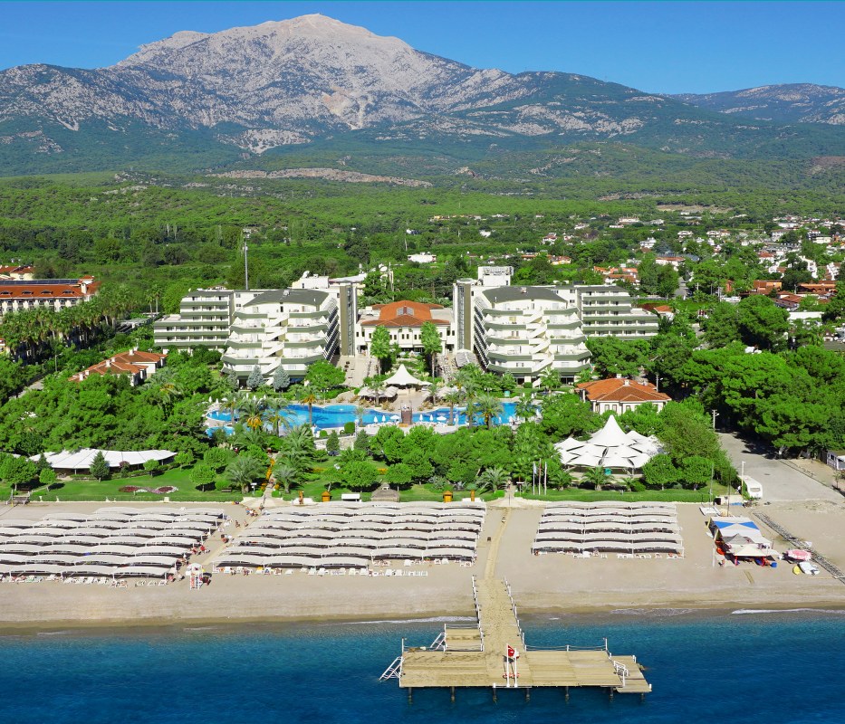 Rai Premium Tekirova skats no jūras puses uz privāto pludmali, piestātni, viesnīcas ēkām un zaļo kalnu ainavu tālumā