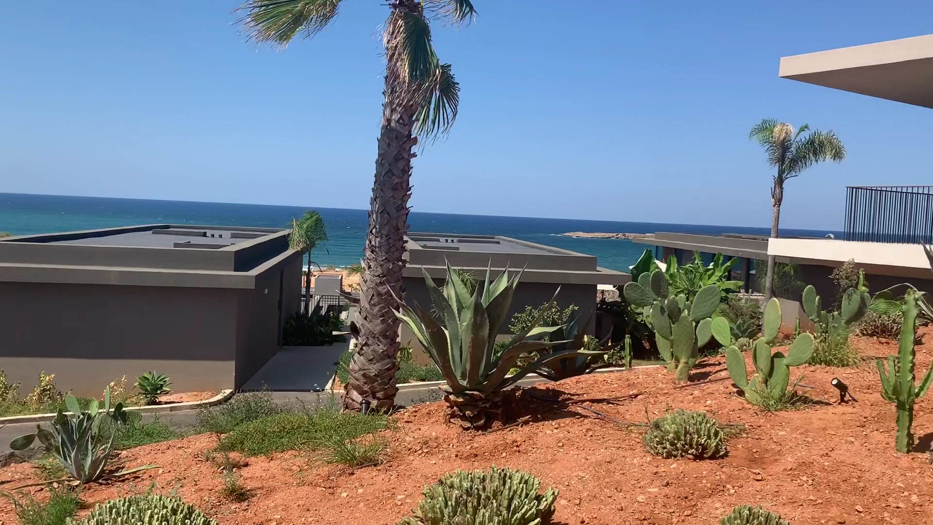 Skats no Domes Zeen Chania dārza uz zilajiem jūras ūdeņiem, priekšplānā kaktusi, agaves un palmas piekrastes kāpās