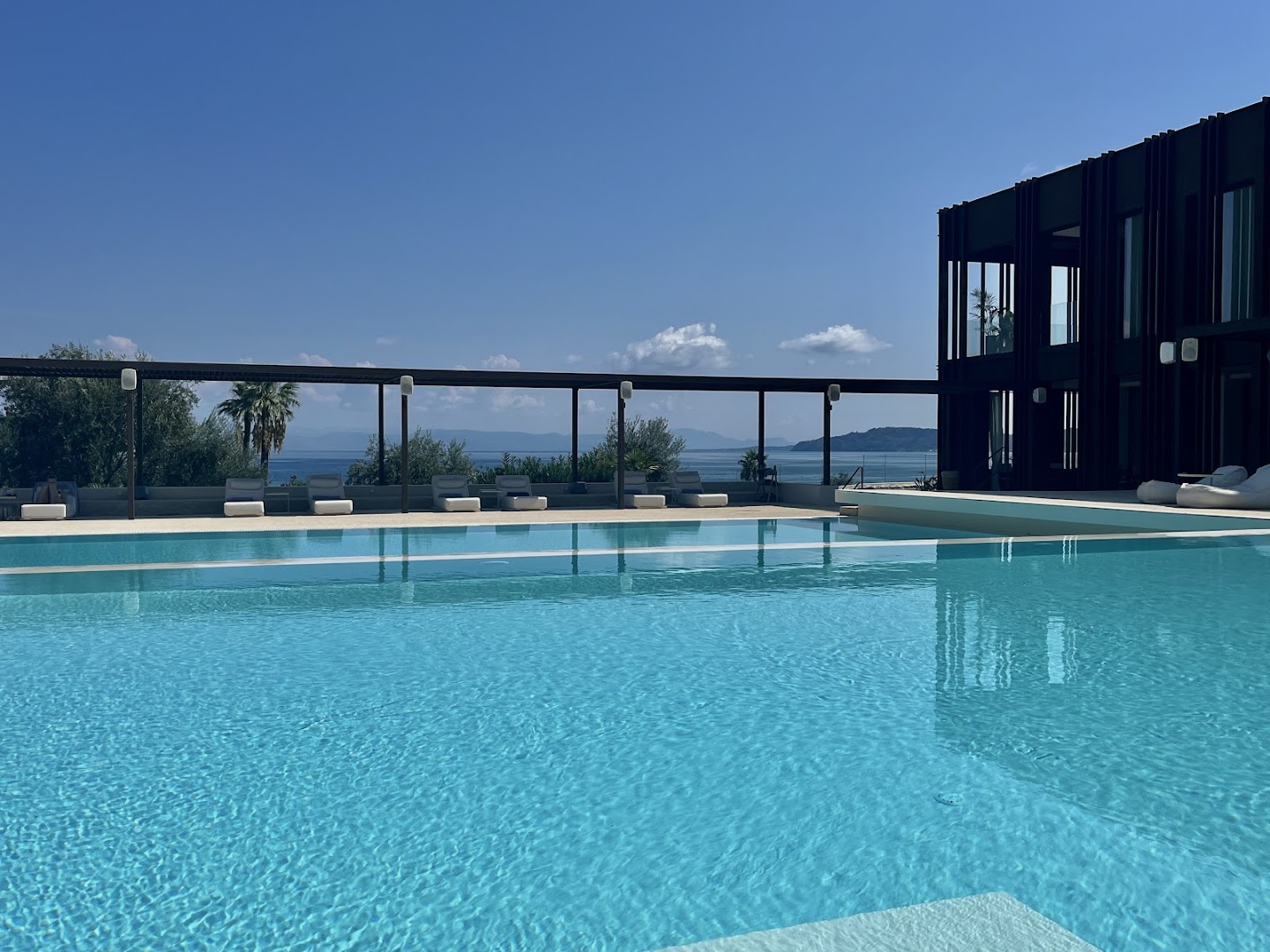 Domes Miramare Resort lielais āra baseins ar skatu uz jūru un kalniem — sauļošanās krēsli zem nojumes Korfu