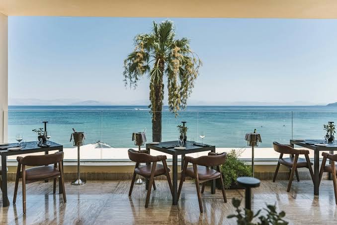Domes Miramare Resort restorāna terase ar skatu uz Jonijas jūru Korfu piekrastē — eleganti galdiņi, koka krēsli un palma saulainā dienā