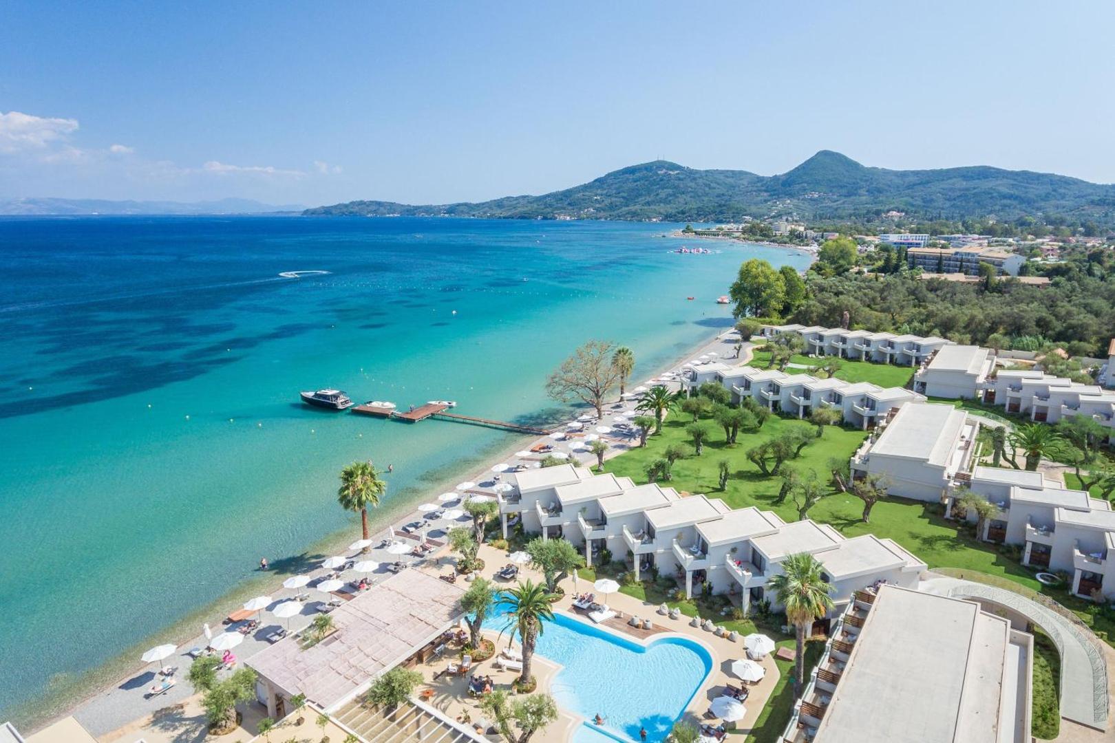 Domes Miramare Resort skats no putna lidojuma uz piekrasti Korfu salā — zilganzaļš jūras ūdens, balta smilšu pludmale un kūrorta bungalo