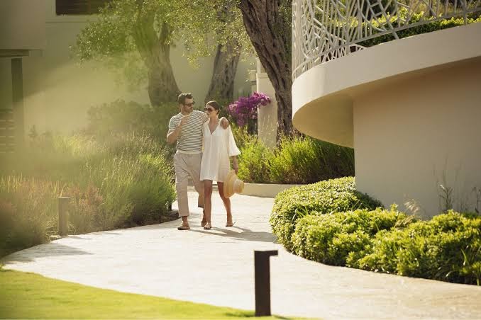 Domes Miramare Resort dārza teritorija ar pāri, kas pastaigājas pa akmens celiņu — zaļojoši augi un moderna arhitektūra Korfu