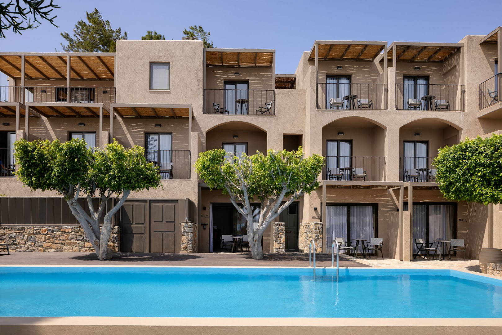 Domes Aulus Elounda Resort viesnīcas korpuss ar numuru balkoniem un baseinu priekšplānā, ko ieskauj koki