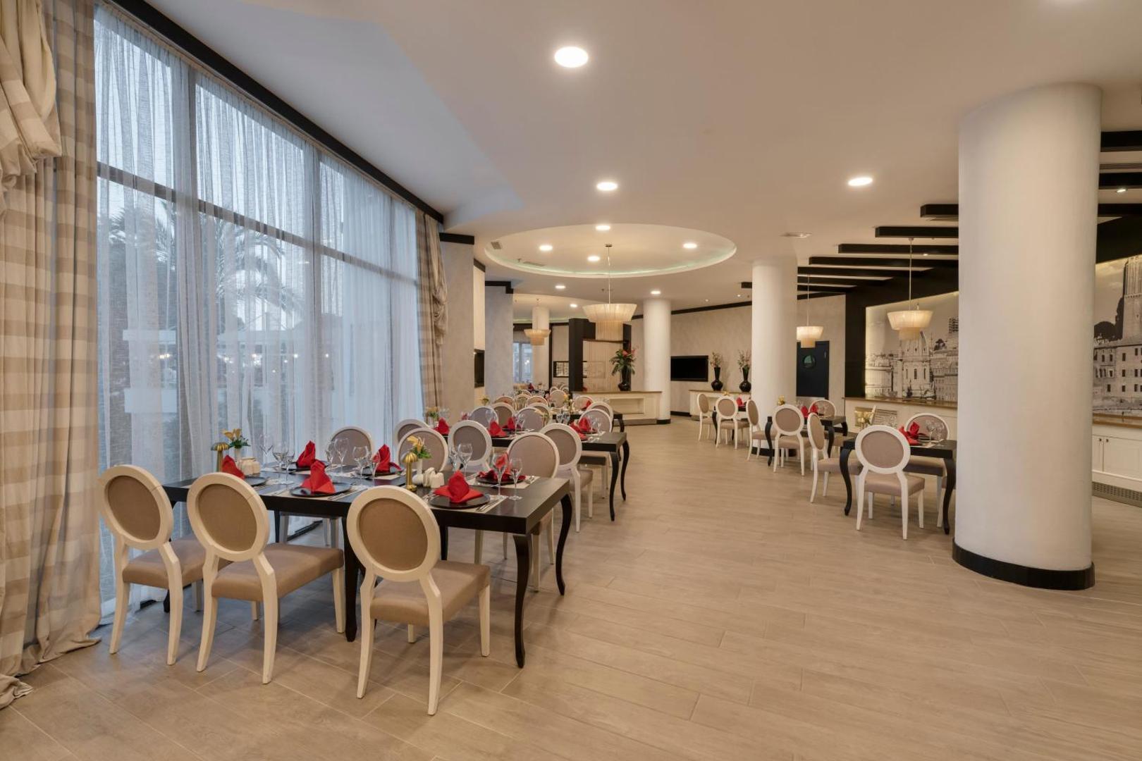 Limak Atlantis De Luxe Hotel & Resort restorāns ar eleganti klātiem galdiem, sarkaniem akcentiem un plašiem logiem uz dārzu Belekā.