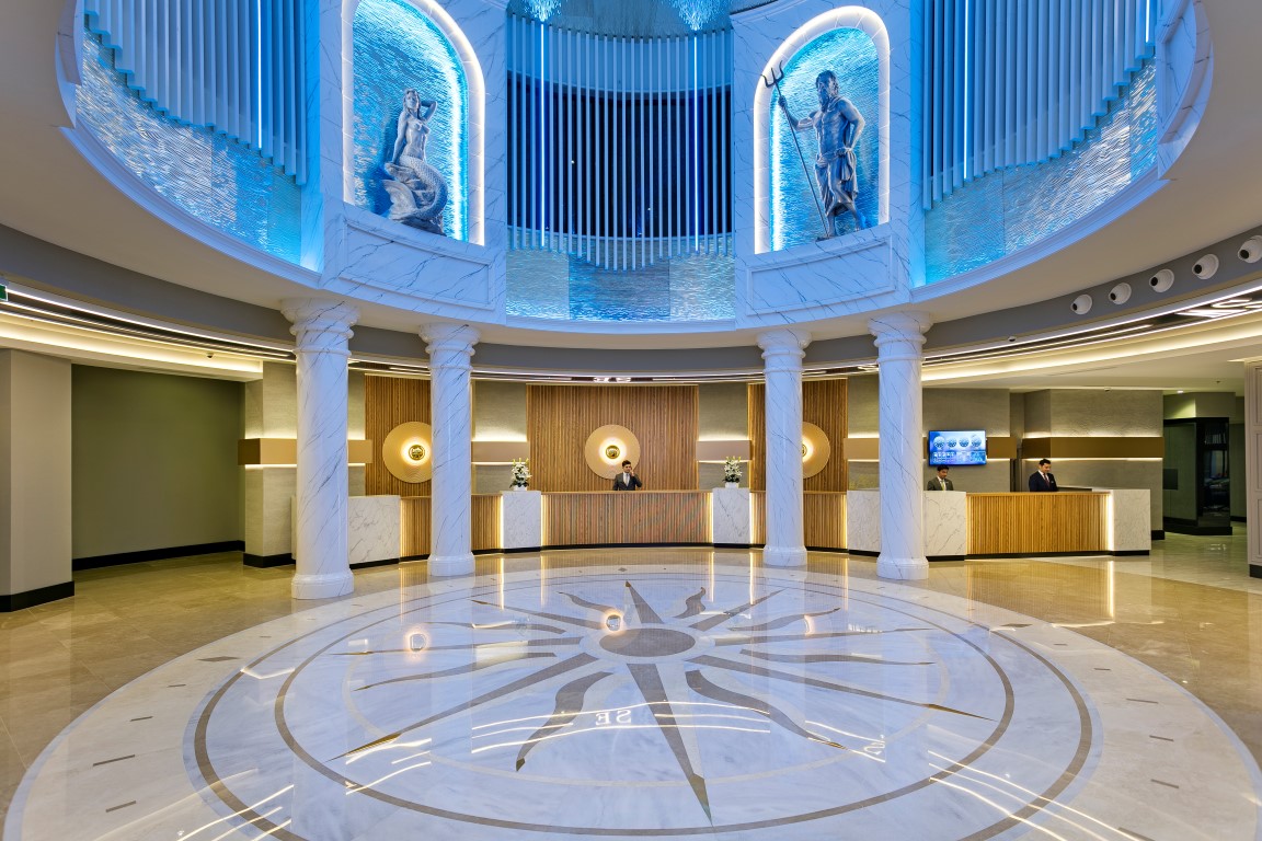 Limak Atlantis De Luxe Hotel & Resort grandiozā recepcijas zāle ar marmora grīdu, kolonnām un antīka stila skulptūrām zilā apgaismojumā.