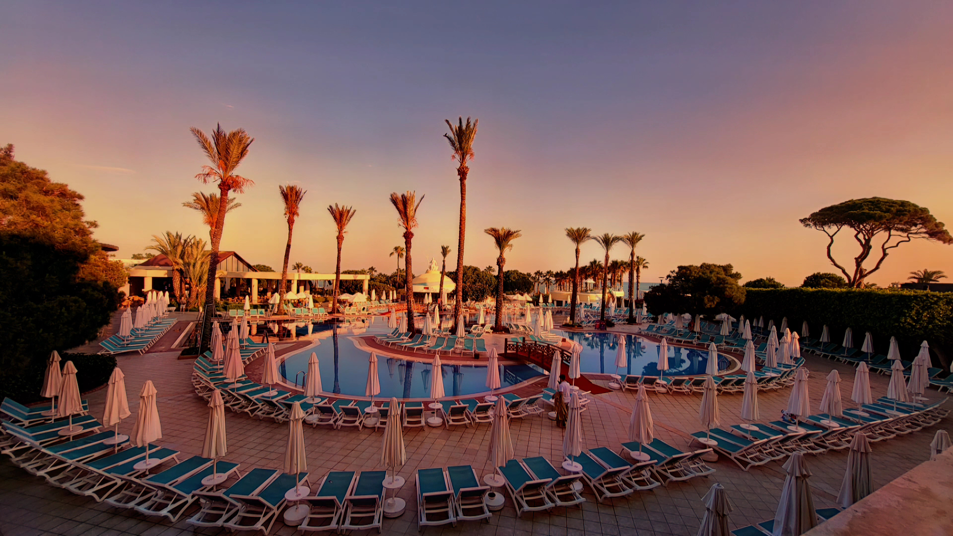 Limak Atlantis De Luxe Hotel & Resort galvenais baseins saulrieta laikā ar palmām un izgaismotu debess jumu Turcijā