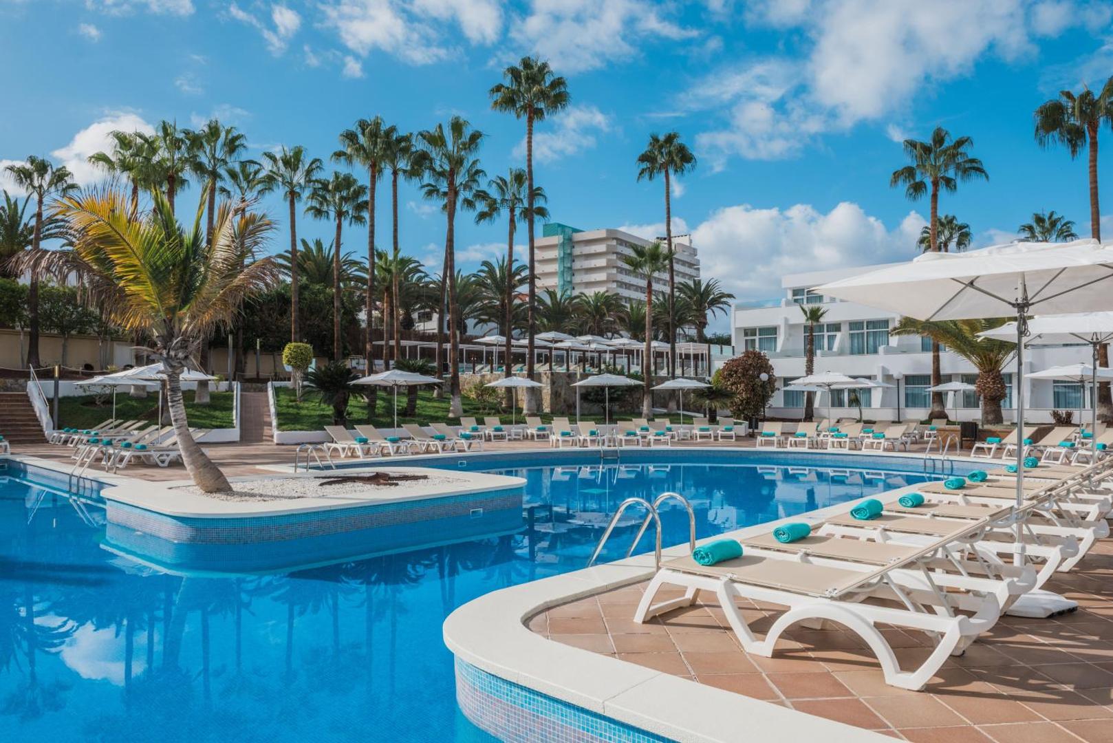 Iberostar Waves Las Dalias āra baseins ar sauļošanās krēsliem un saulessargiem Tenerifē — plaša atpūtas zona ar palmām un zilu debesi
