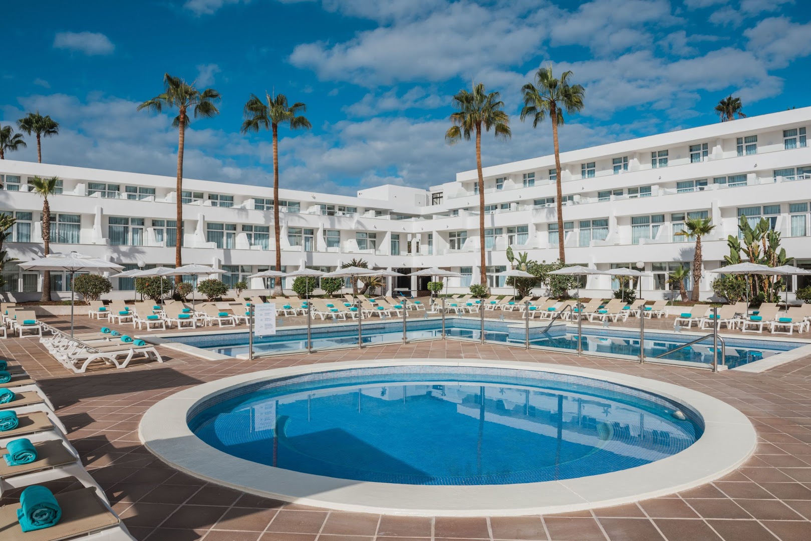Iberostar Waves Las Dalias bērnu baseins un sauļošanās zona — gaiša viesnīcas arhitektūra, palmas un zilas debesis fonā