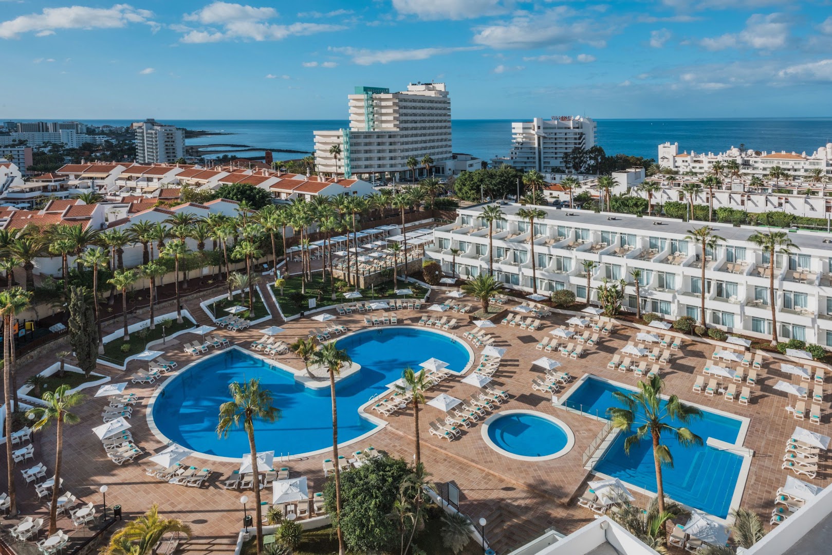 Iberostar Waves Las Dalias āra baseini un sauļošanās terase Tenerifē — gaiša viesnīcas arhitektūra, palmas un debeszils ūdens