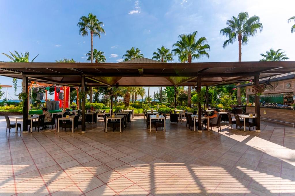 L'ancora Beach Hotel āra restorāna terase zem nojumes ar skatu uz dārzu un palmām Turcijas piekrastē saulainā dienā.