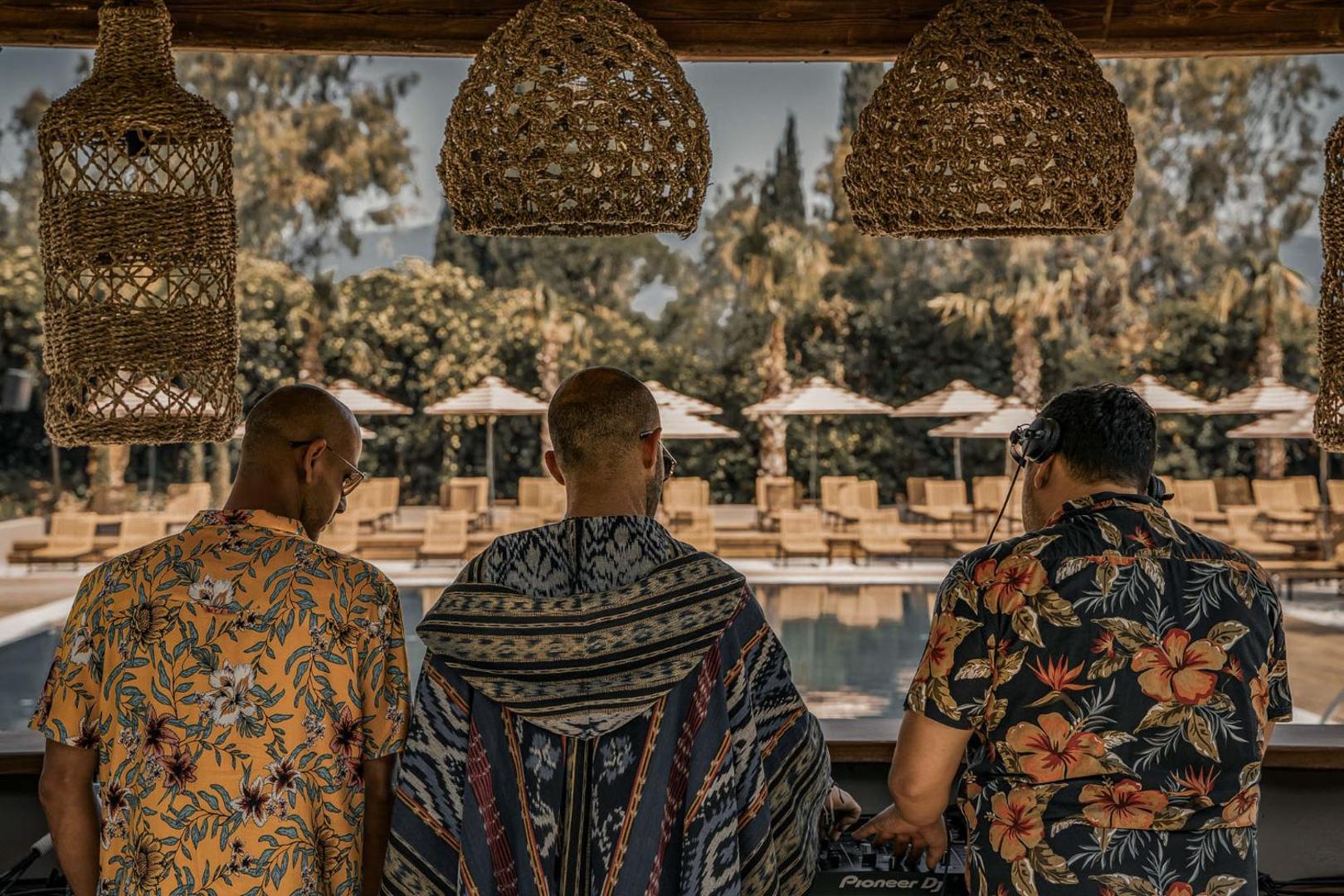 Cook's Club Corfu dīdžeji pie pults pie āra baseina, fonā saulessargi un palmas Grieķijas kūrortā