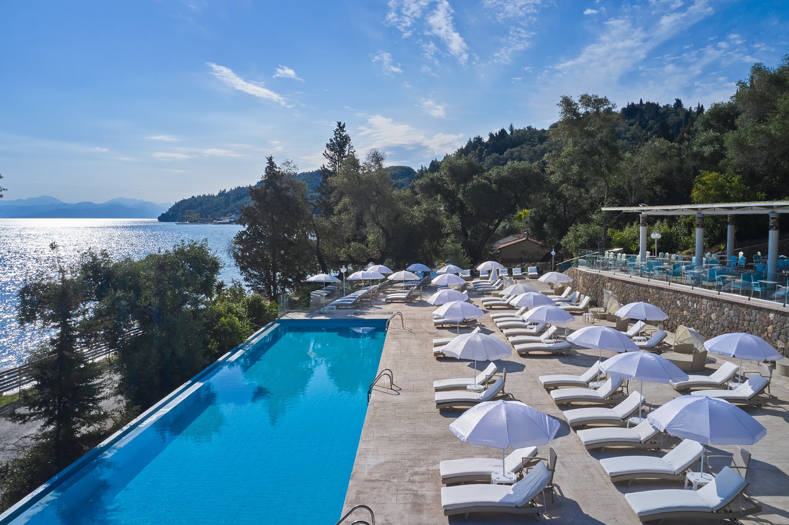 Mythos Palace Resort āra baseins ar pārplūdes malu un baltiem saulessargiem Jonijas jūras krastā