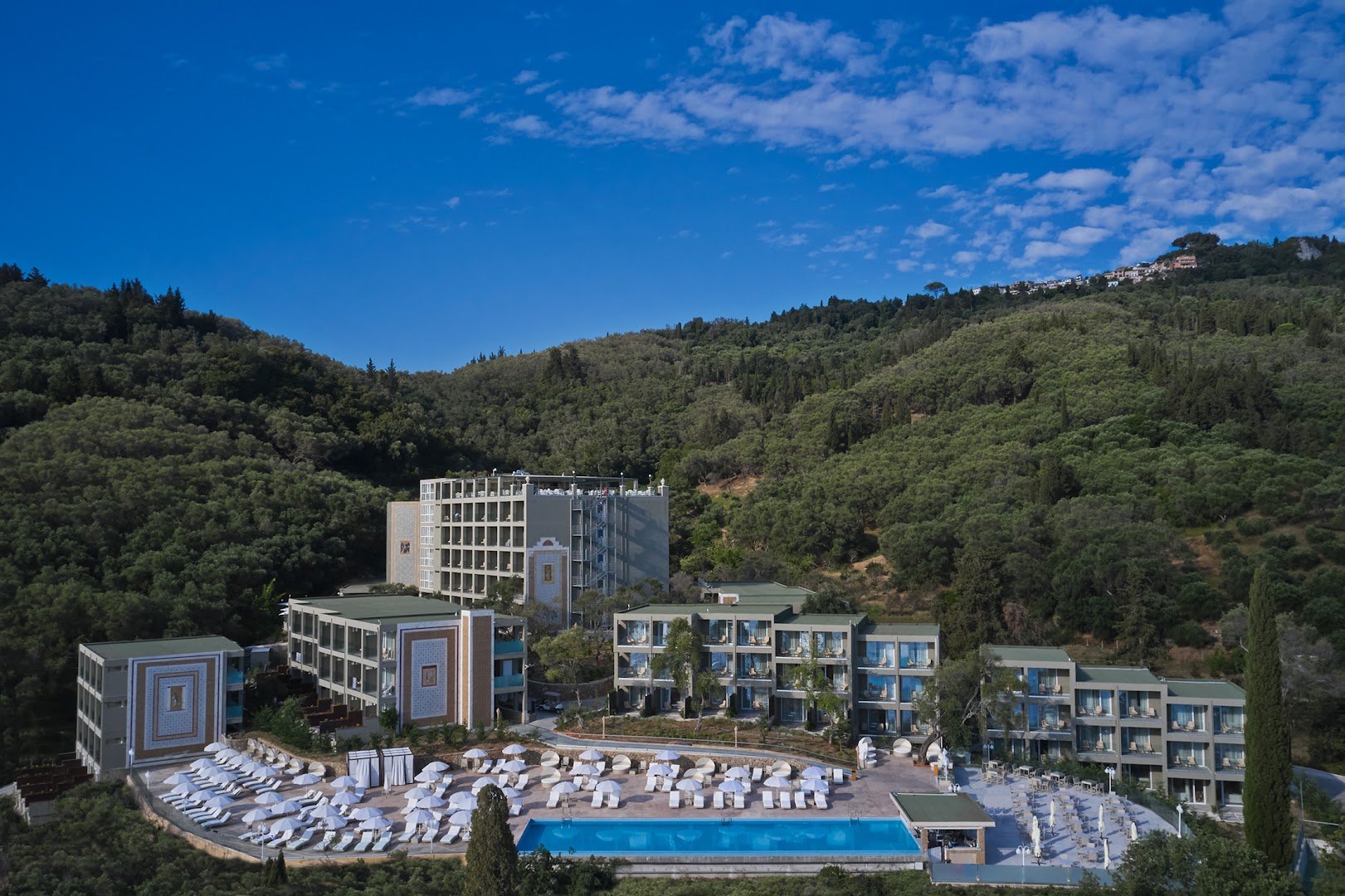 Mythos Palace Resort ēku komplekss ar āra baseinu un saulessargiem Korfu piekrastē, ieskauts olīvkoku birzīs un kalnos