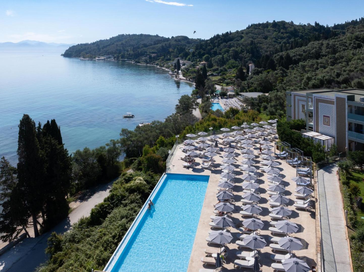 Mythos Palace Resort teritorijas skats no augšas ar baseinu, saulessargiem un piekrastes līniju Korfu