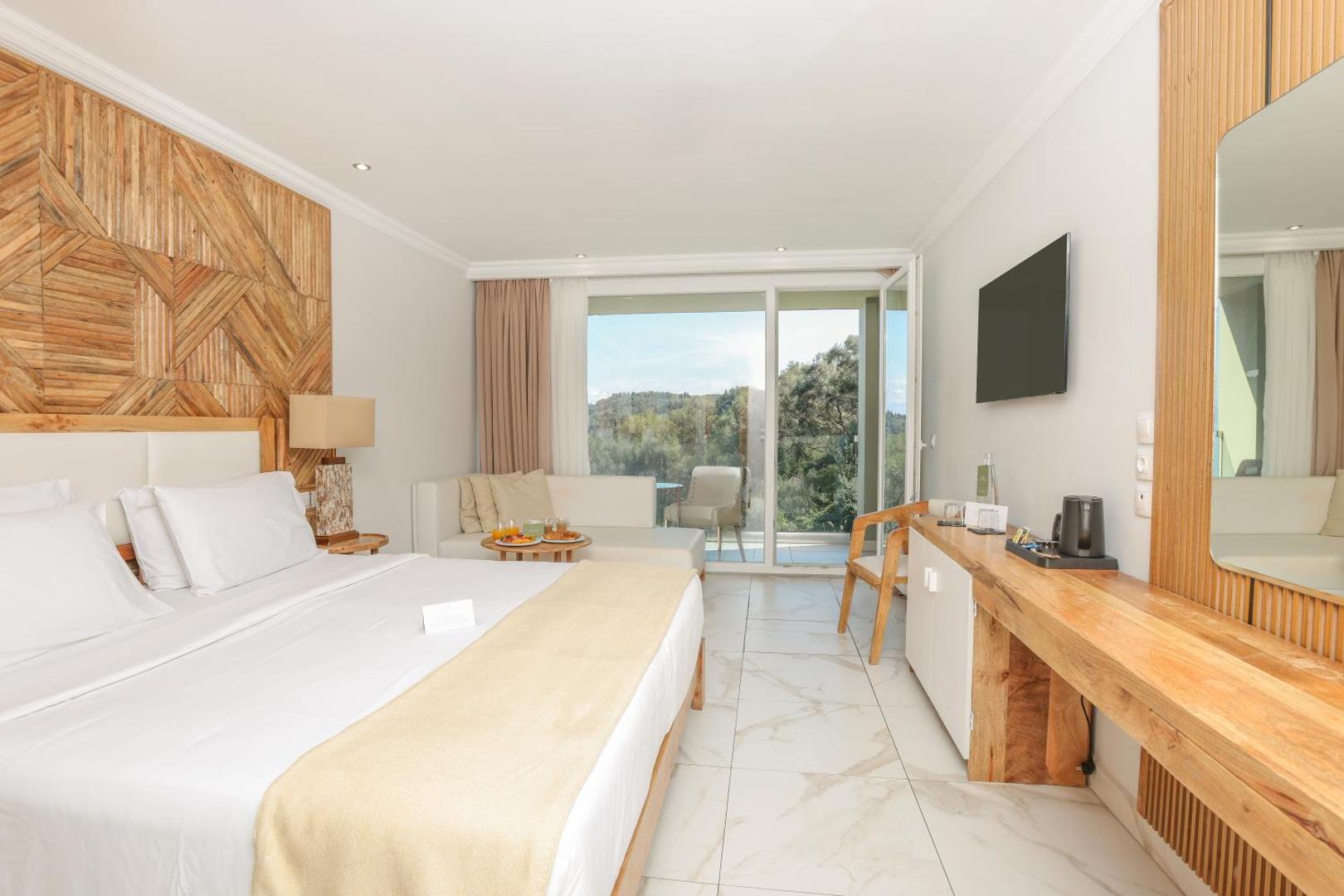 Gaišs un mājīgs Mythos Palace Resort viesnīcas numurs ar balkonu, no kura paveras skats uz Korfu zaļajiem kalniem