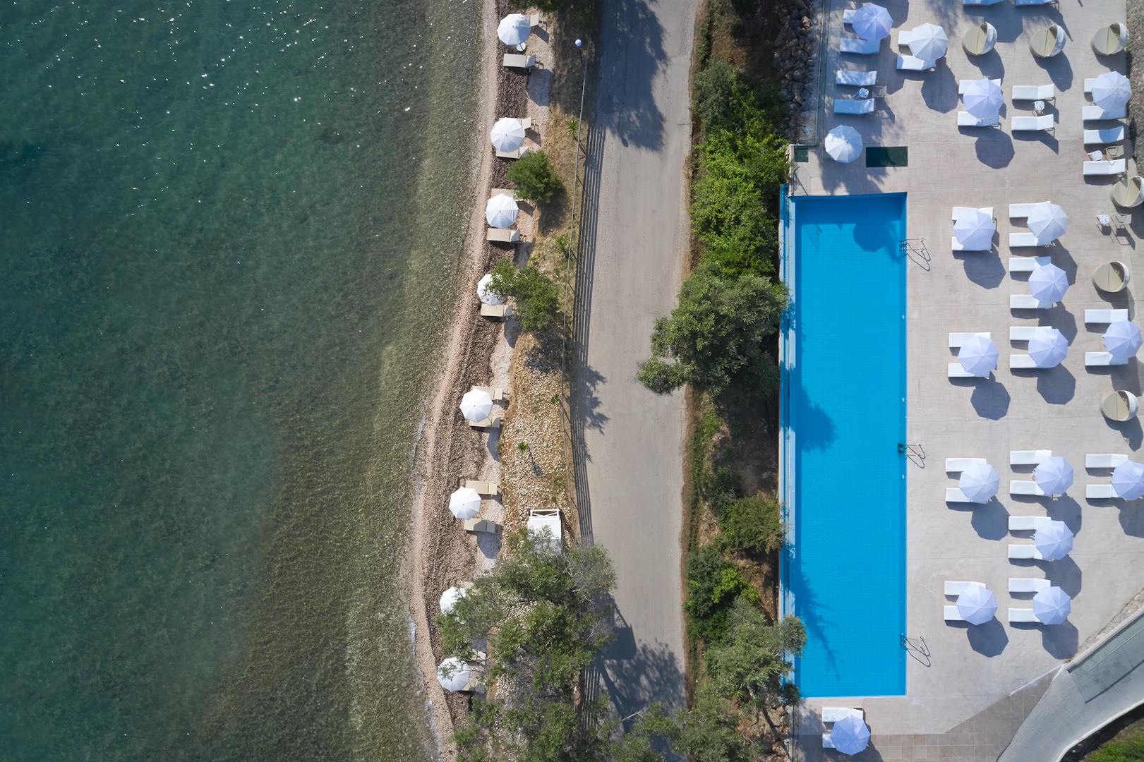 Aerofoto skats uz Mythos Palace Resort baseinu un blakus esošo oļu pludmali ar baltiem saulessargiem Korfu salā