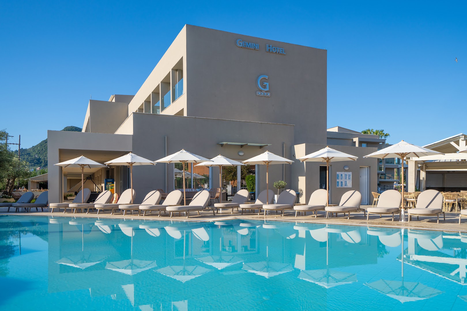 CNic Gemini Hotel atpūtas krēsli un saulessargi pie baseina Mesongi kūrortā Korfu — sakopta vide un moderna viesnīcas arhitektūra