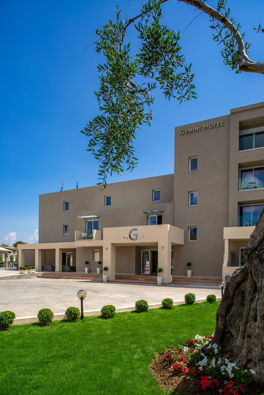 CNic Gemini Hotel fasāde un ieeja Mesongi ciemā Korfu salā — skats no dārza ar zālāju un olīvkoku saulainā dienā