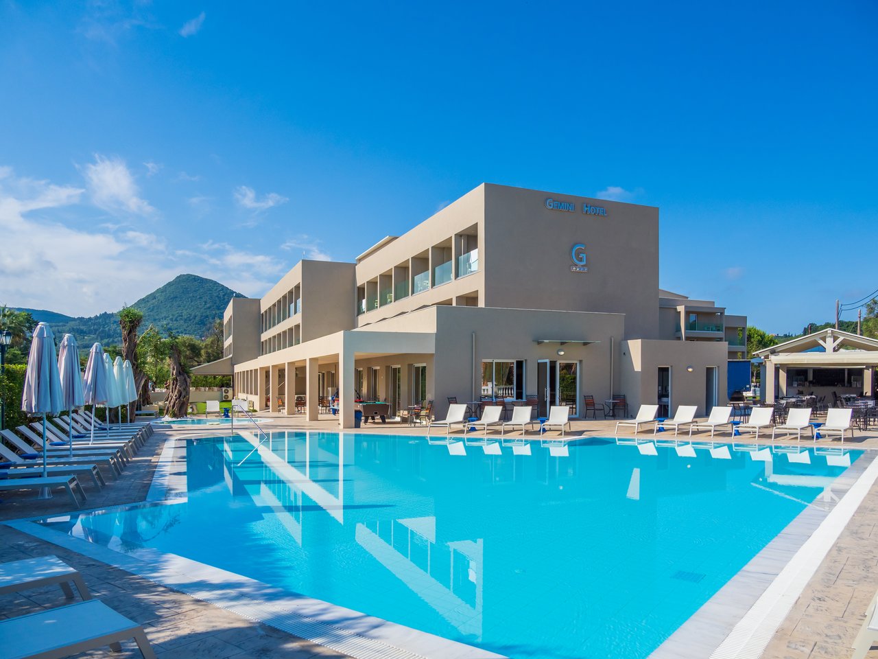 CNic Gemini Hotel galvenā ēka un lielais peldbaseins ar saulessargiem Mesongi, Korfu — plaša sauļošanās terase un skats uz zaļajiem kalniem
