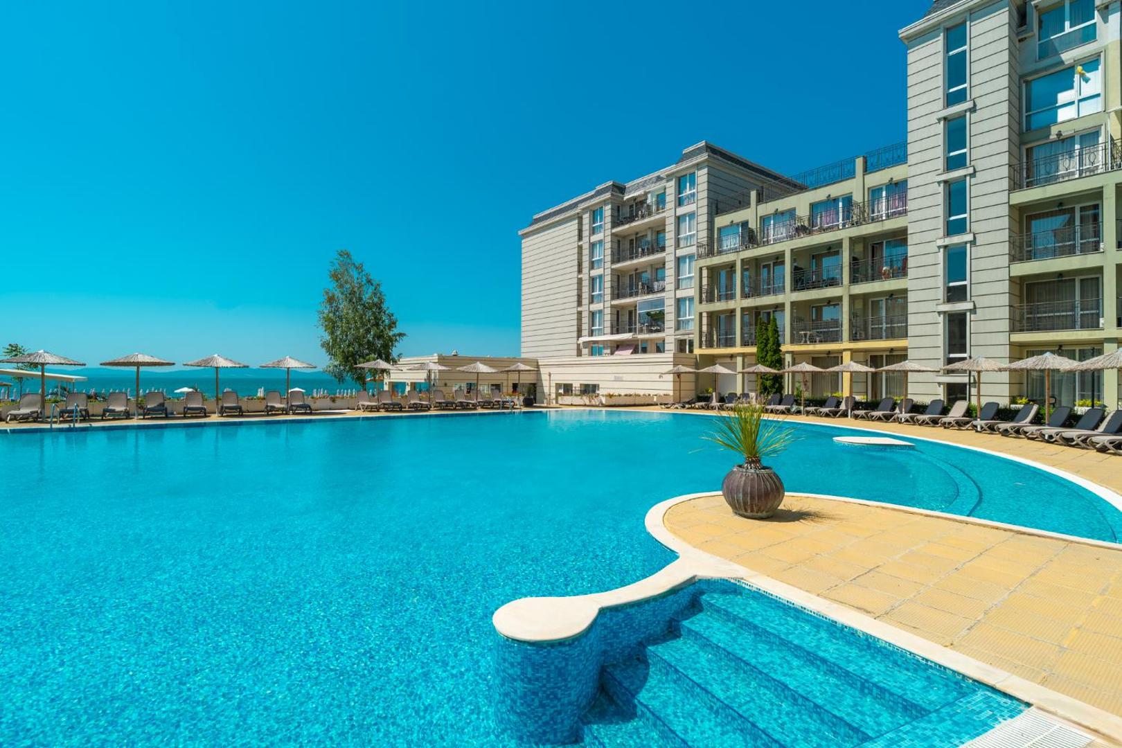 Festa Pomorie Resort lielais āra baseins ar dzidru ūdeni, sauļošanās krēsliem un skatu uz Melno jūru saulainā dienā.