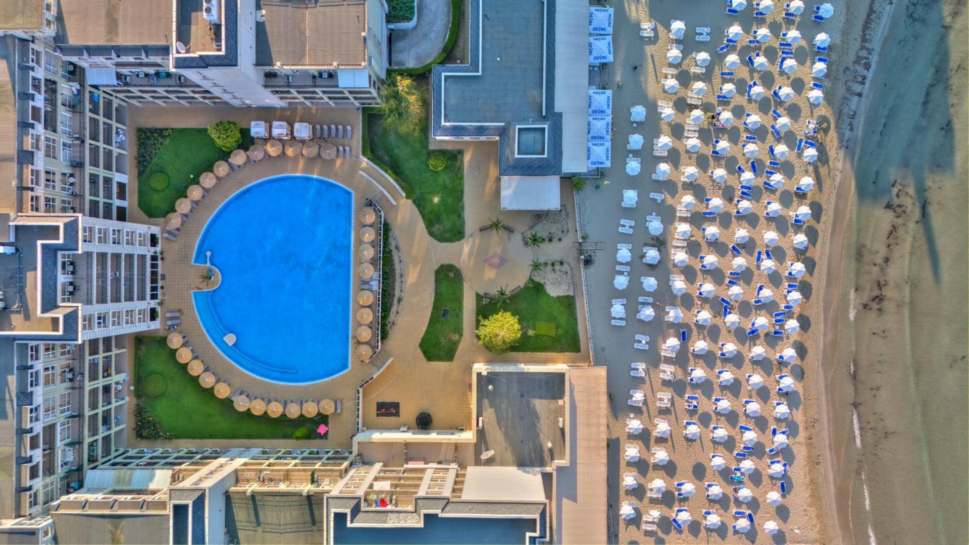 Festa Pomorie Resort skats no putna lidojuma uz apaļo baseinu, viesnīcas korpusiem un plašo smilšu pludmali ar saulessargiem Pomorijā.