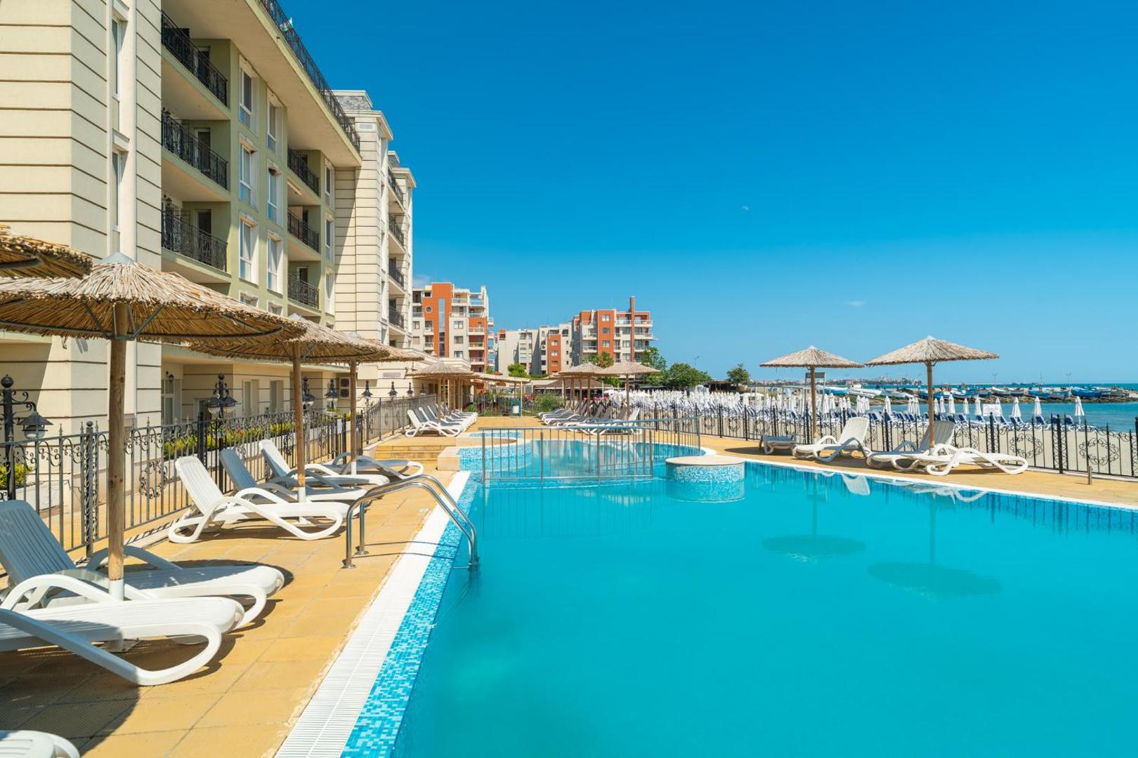 Festa Pomorie Resort baseina zona ar ērtiem zviļņiem un salmu saulessargiem, fonā redzama pludmale un kūrorta apbūve.