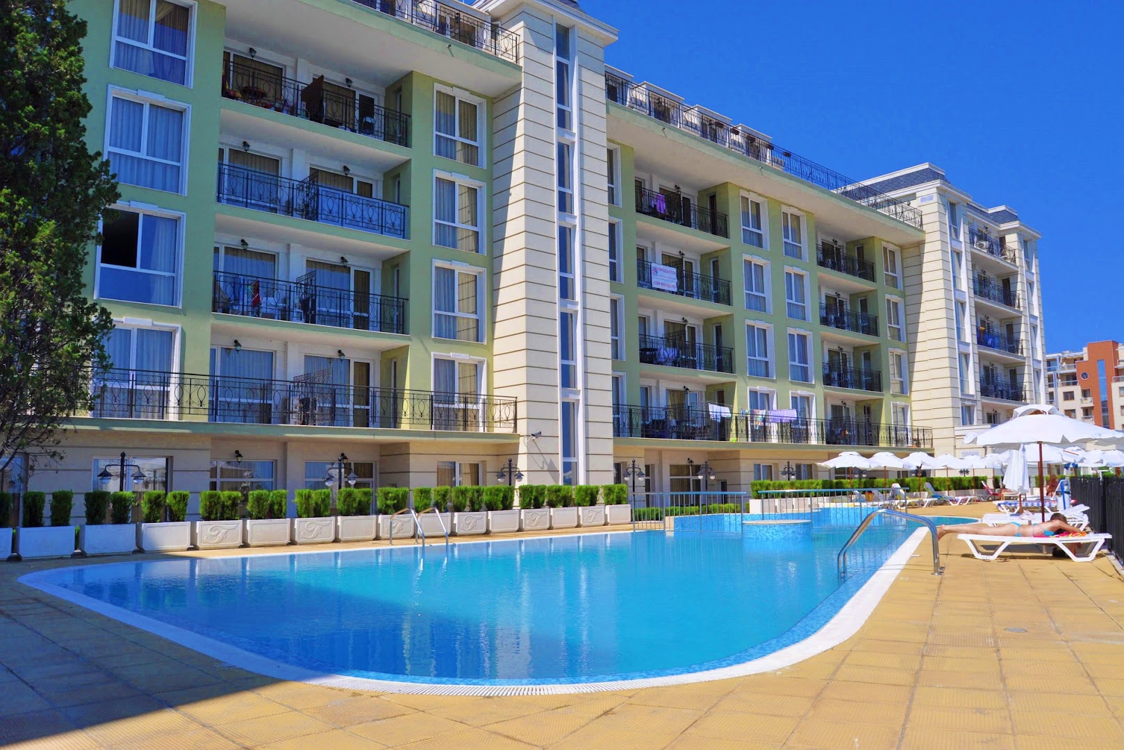 Festa Pomorie Resort daudzstāvu ēkas fasāde ar balkoniem un āra baseinu priekšplānā zem skaidrām, zilām debesīm.