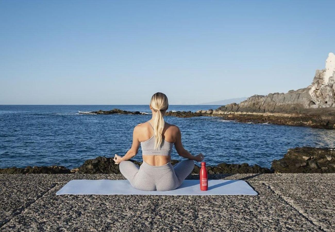 Sieviete meditē uz jogas paklājiņa okeāna krastā pie MYND Adeje viesnīcas Tenerifē, fonā zilais ūdens un klintis