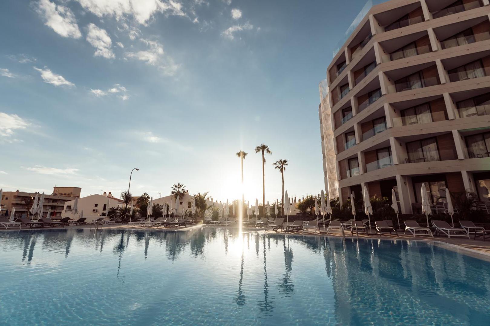 Viesnīcas MYND Adeje modernā ēka un baseins saulrietā ar palmām un saulessargiem Tenerifes kūrortā