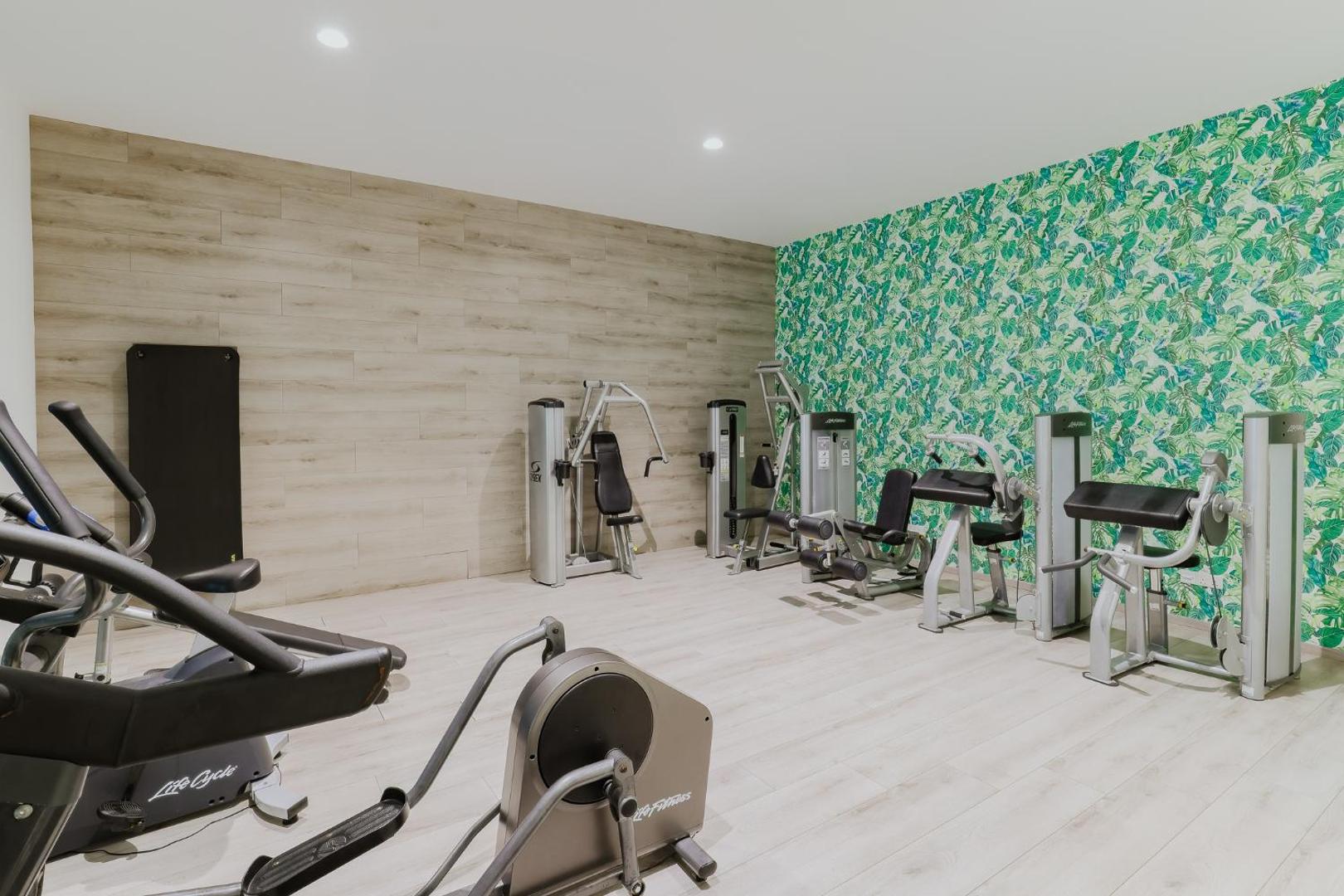 Tasia Maris Seasons Hotel modernā trenažieru zāle ar Life Fitness aprīkojumu — krosa trenažieri un spēka iekārtas telpā ar tropisku tapešu dizainu.