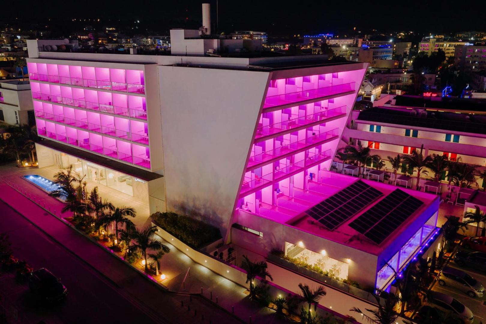 Tasia Maris Seasons Hotel ēka naktī ar koši rozā balkonu apgaismojumu Ajanapā, Kiprā — moderna arhitektūra un pilsētas panorāma