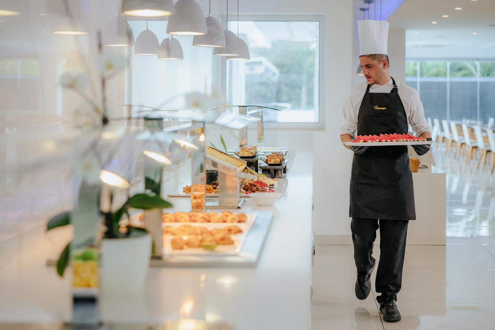 Tasia Maris Seasons Hotel pavārs baltā cepurē pasniedz svaigus augļus pie restorāna bufetes galda ar uzkodām un konditoreju