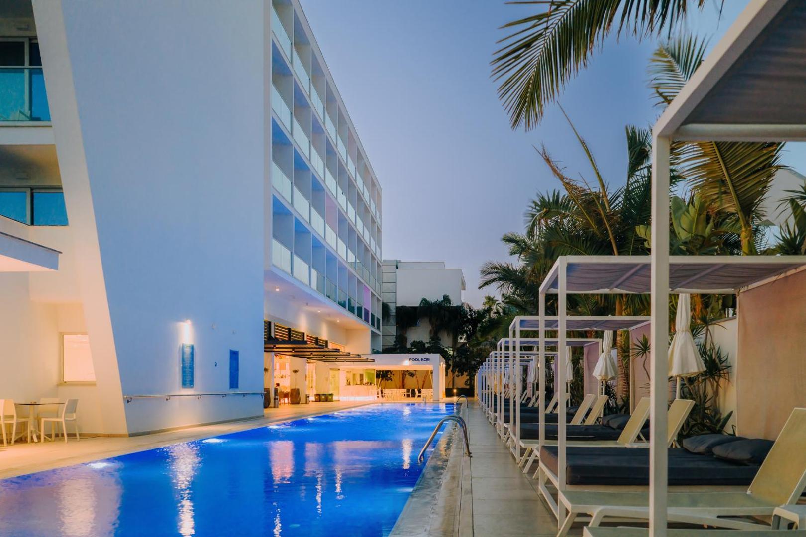 Tasia Maris Seasons Hotel izgaismots āra baseins vakarā ar atpūtas nojumēm un palmām Aijanapā, Kiprā.