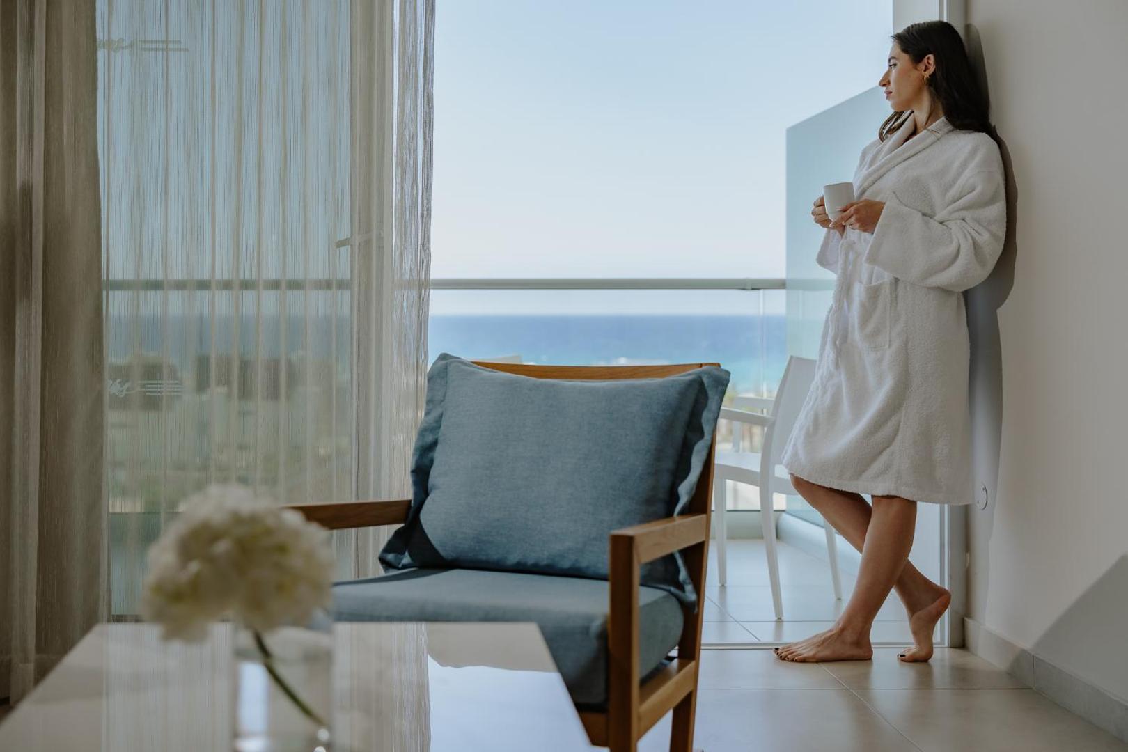 Sieviete baltā peldmētelī uz Tasia Maris Seasons Hotel numura balkona ar skatu uz Vidusjūru Aijanapā.