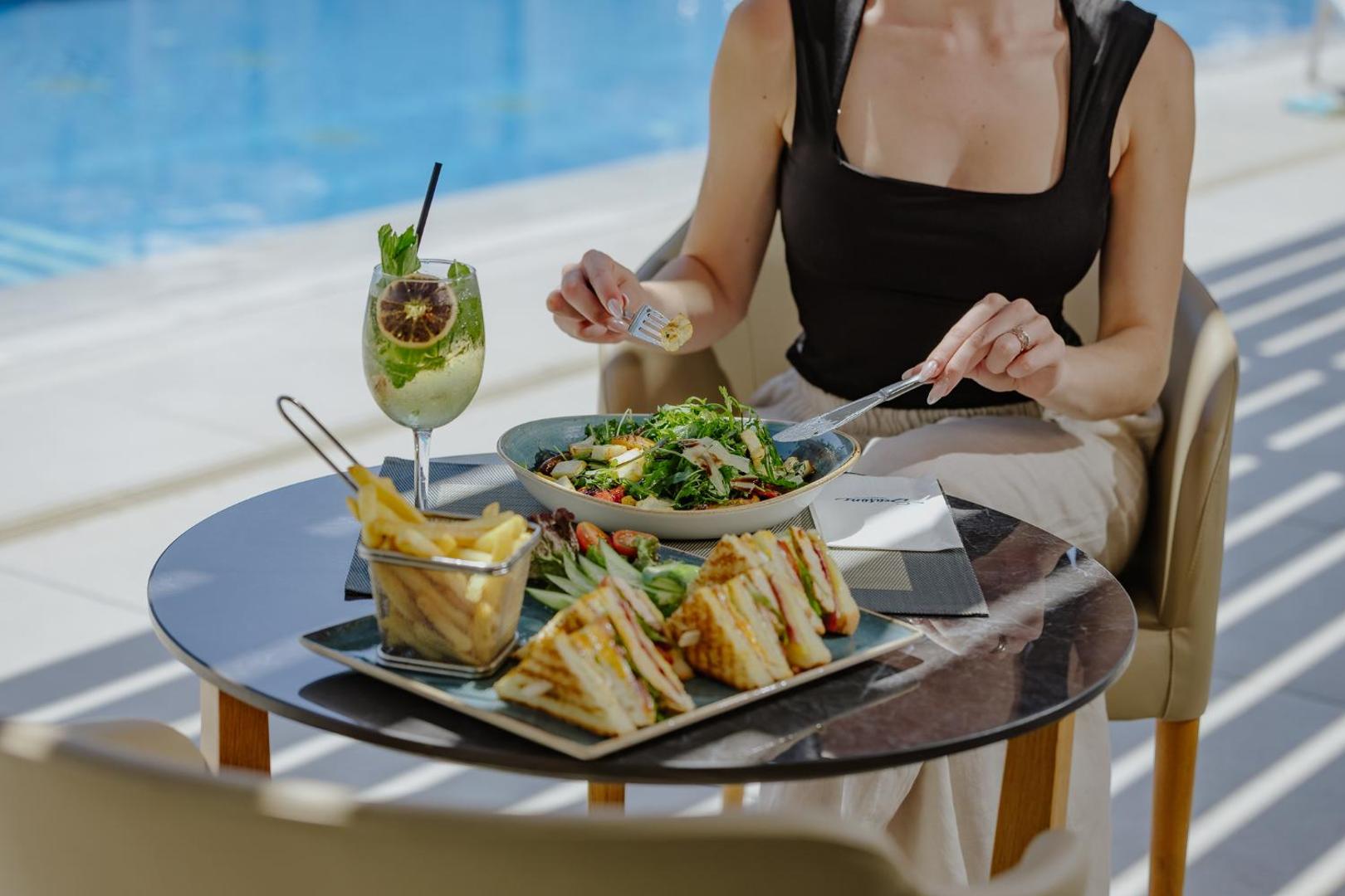 Tasia Maris Seasons Hotel pusdienas pie baseina Kiprā — sieviete bauda svaigus salātus, klubu sviestmaizi un kokteili saulainā āra terasē.