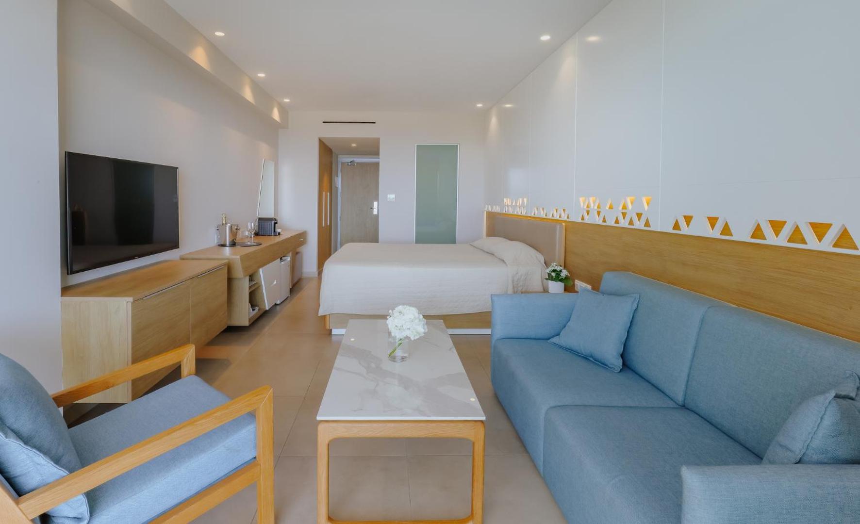 Tasia Maris Seasons Hotel plašs un gaišs viesnīcas numurs ar divguļamo gultu, dīvānu un televizoru Aijanapas kūrortā.