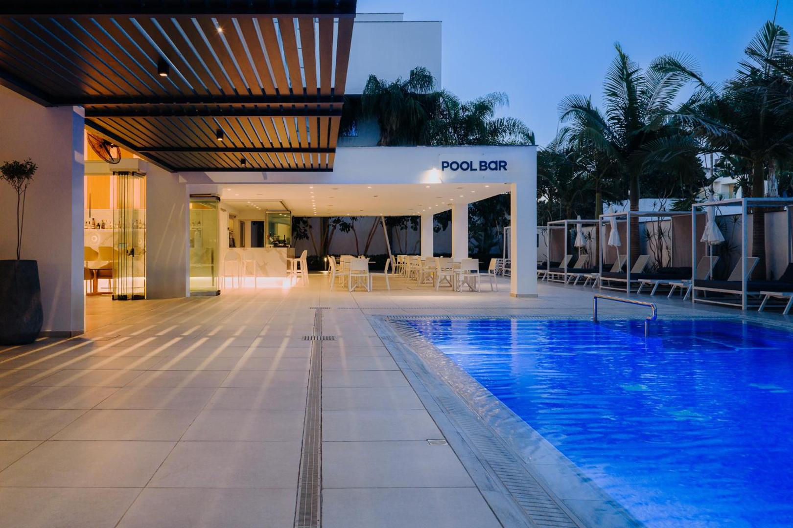 Tasia Maris Seasons Hotel āra baseins un Pool Bar vakara apgaismojumā Ajanapā, Kiprā — moderna atpūtas zona ar galdiņiem un palmām