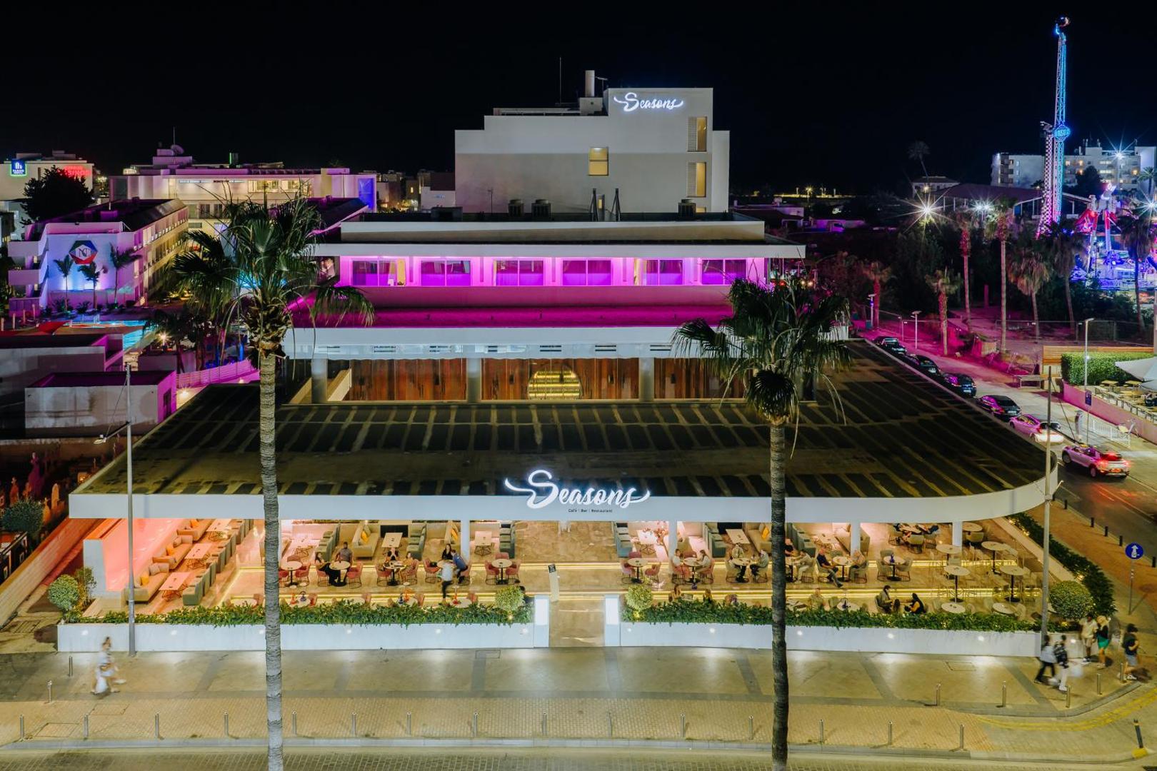 Tasia Maris Seasons Hotel galvenā ēka un restorāns naktī ar košu neona apgaismojumu un āra terasi Ajanapas centrā