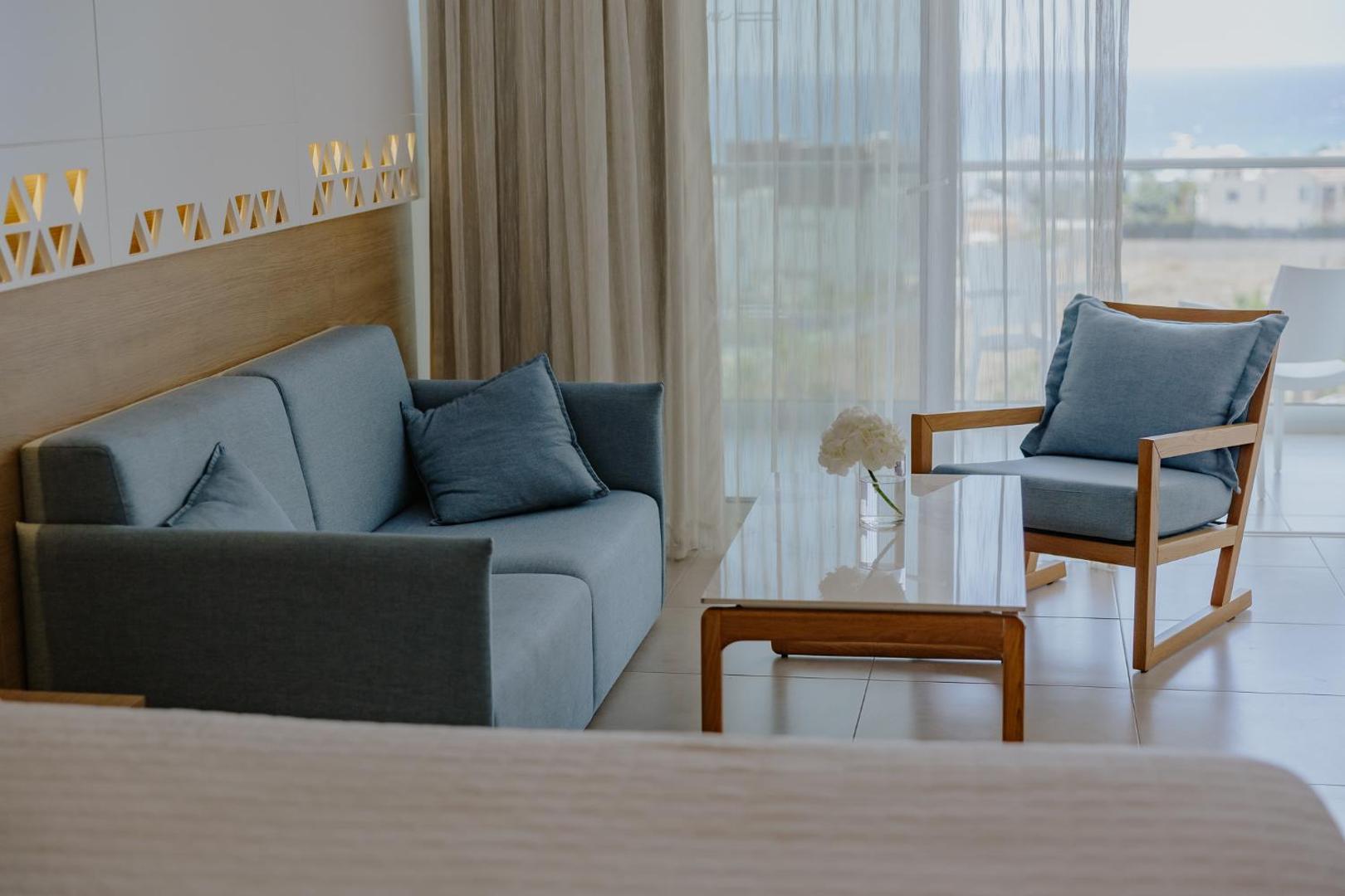 Tasia Maris Seasons Hotel numura atpūtas zona ar gaiši zilu dīvānu, koka atpūtas krēslu un kafijas galdiņu Aijanapā, Kiprā.