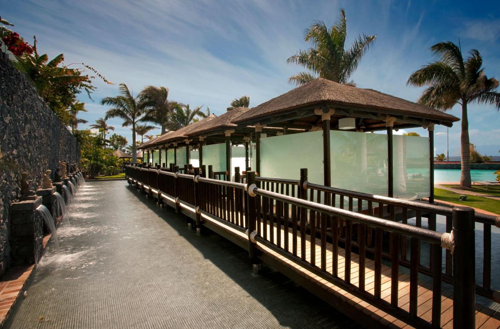 Gran Melia Palacio de Isora spa zonas āra paviljoni ar salmu jumtiem pie dekoratīva ūdens baseina un palmām