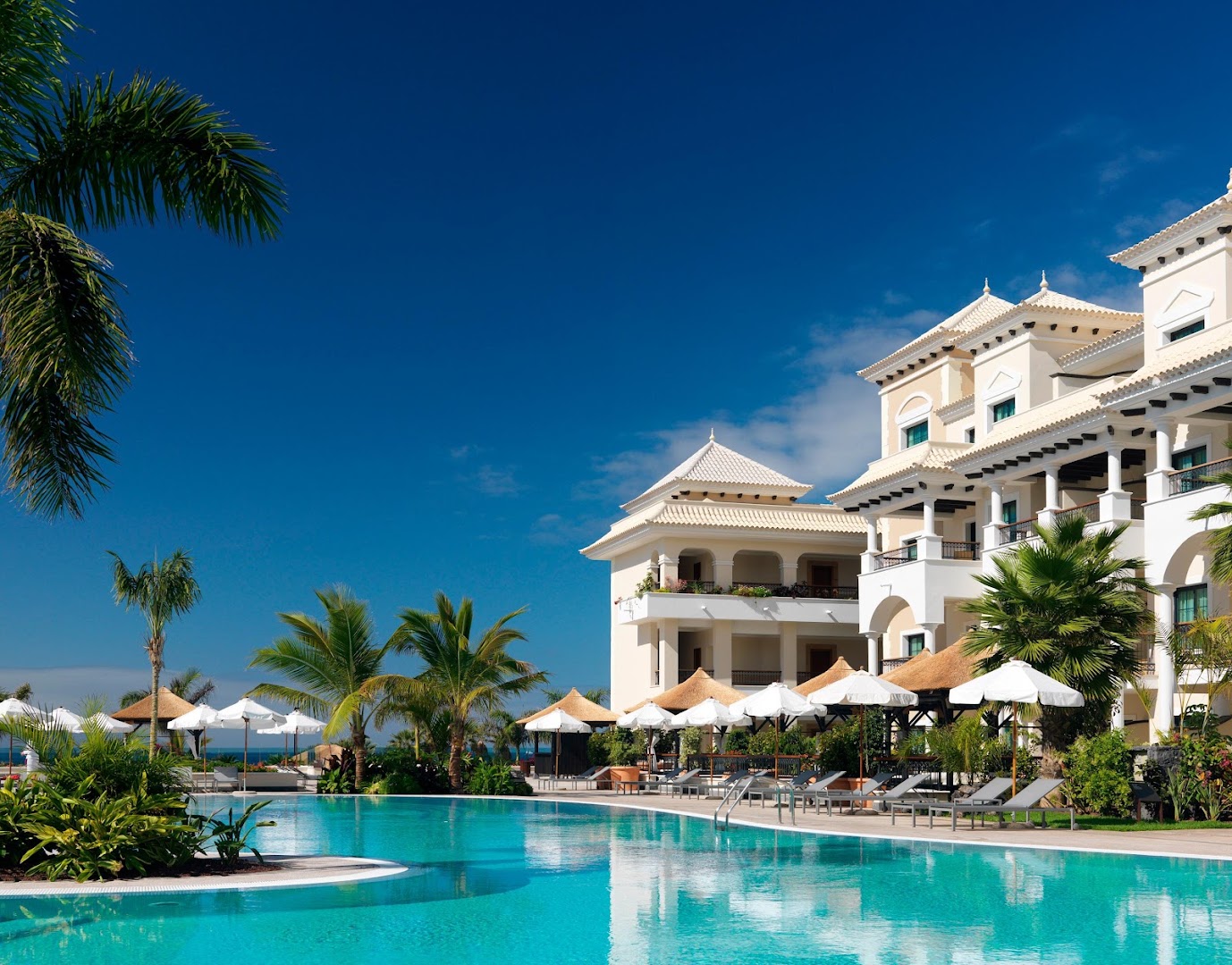Gran Melia Palacio de Isora āra baseins Tenerifē — grezna viesnīcas fasāde, sauļošanās krēsli ar saulessargiem un palmas zilās debesīs