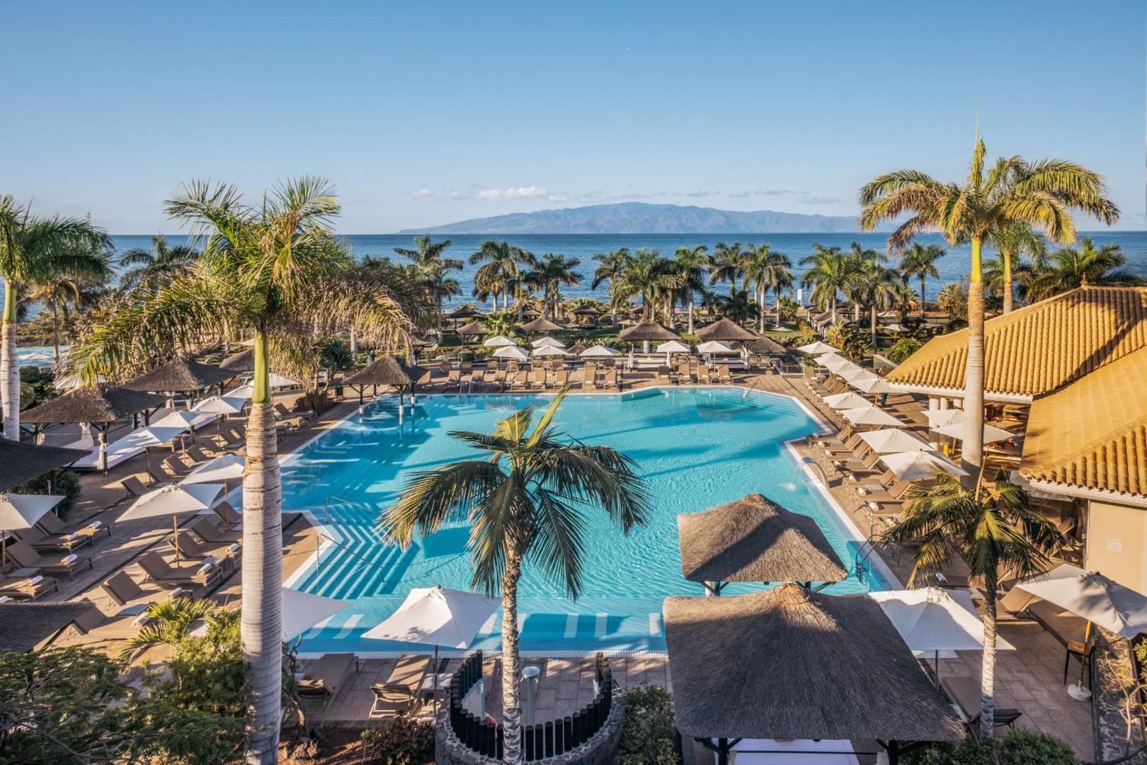 Gran Melia Palacio de Isora panorāmas skats uz galveno baseinu, palmām un okeānu Tenerifē no augšējā stāva terases.