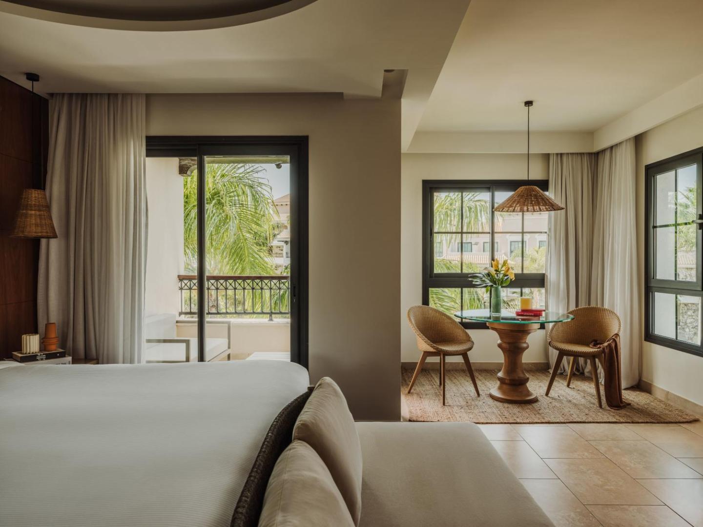 Gran Melia Palacio de Isora gaišs viesnīcas numurs ar atpūtas zonu, pītiem krēsliem un skatu uz zaļojošu dārzu caur balkona durvīm