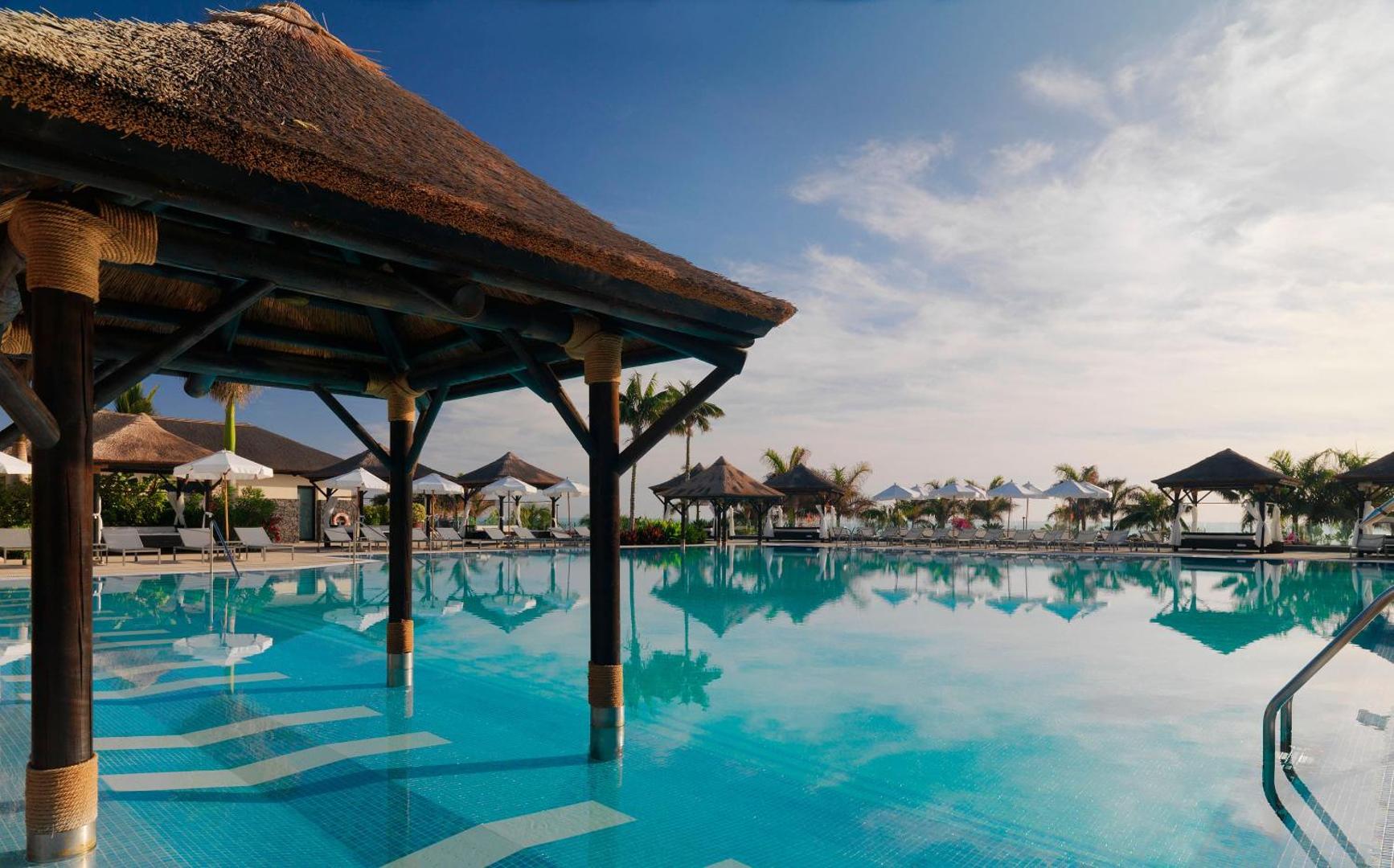 Gran Melia Palacio de Isora plašais āra baseins ar atpūtas nojumēm un saulessargiem Tenerifes kūrortā pie okeāna.